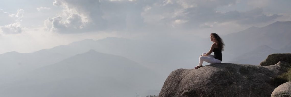 A woman enjoys serene solitude, sitting on a mountain rock with a vast view of the sunrise landscape.